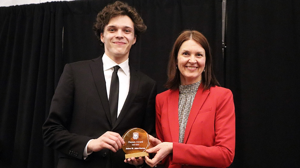 A man and woman stand side by side, displaying an award in front of a dark black background.